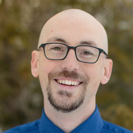 zach - WPChat Smiling bald man with glasses wearing a blue shirt, outdoors, in focus with a natural background.